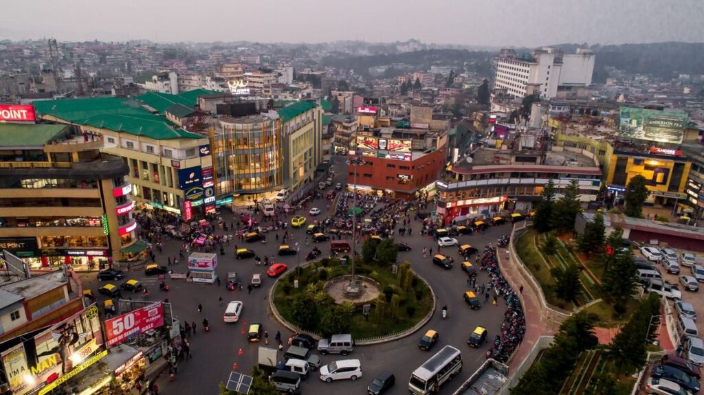 Busy Shillong city Centre, with crowded roundabouts, taxis, pedestrians, shops, and green rooftops reflecting lively urban life.