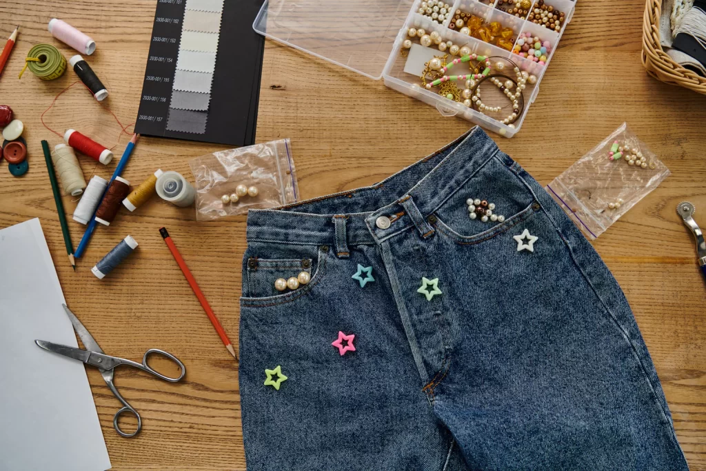 Denim jeans decorated with pearls and star buttons, surrounded by sewing tools and threads on a table.
